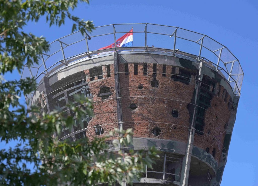 VUKOVAR: GRAD KOJI JE PROŠAO KROZ PAKAO, ALI JE 2025. GODINE OPET USPRAVNO STAO PRED HRVATSKU Vukovarski vodotoranj, simbol otpora, prikazan izbliza s vidljivim oštećenjima od granata, okružen skelama i zaštitnom mrežom, s hrvatskom zastavom na vrhu i plavim nebom u pozadini, uokviren granama drveća.